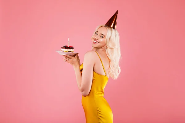 正若いですブロンド女性でドレスとパーティー帽子保持ピースのケーキでキャンドルと笑顔でカメラ ポーズピンクのスタジオの背景に孤立 フリースペース — ストック写真