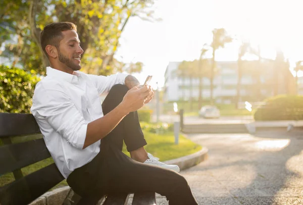 携帯電話や笑顔を使用して正式な衣装で陽気な若いビジネスマン 公園でベンチに座って オンラインアシスタントをチェック モバイルアプリ経由でチャット フリースペース — ストック写真