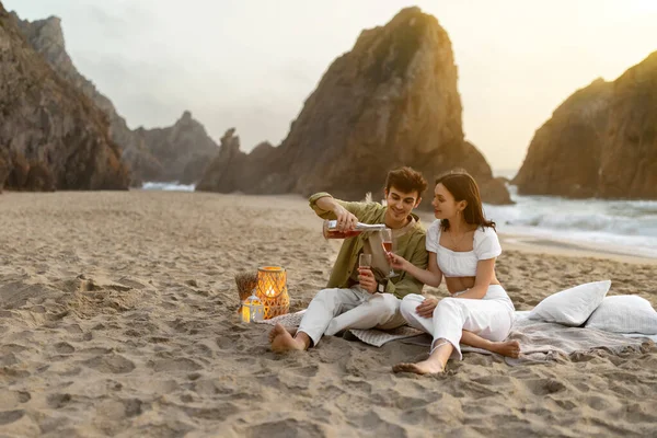 爱你的欧洲夫妻开瓶美酒 在靠近大海的海滩上举行浪漫的野餐 坐在沙滩上的毛毯上享受约会 自由自在 — 图库照片