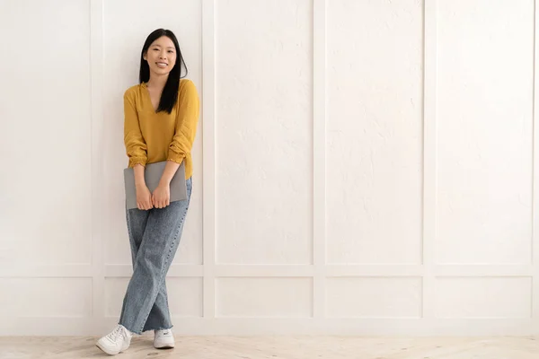 フリーランス リモートジョブ 彼女の手にラップトップで空白の壁の上にポーズカジュアル服のフリーナーを身に着けている幸せな陽気なかなり若いアジアの女性 自宅から働く中国人女性 広告のためのスペースをコピー — ストック写真