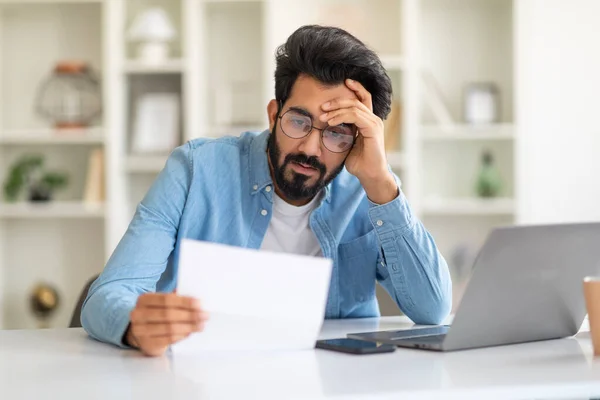 stock image Upset Young Indian Man Sitting At Desk With Laptop And Checking Documents, Focused Millennial Eastern Male Reading Papers While Working Remotely At Home Office, Closeup Shot With Free Space