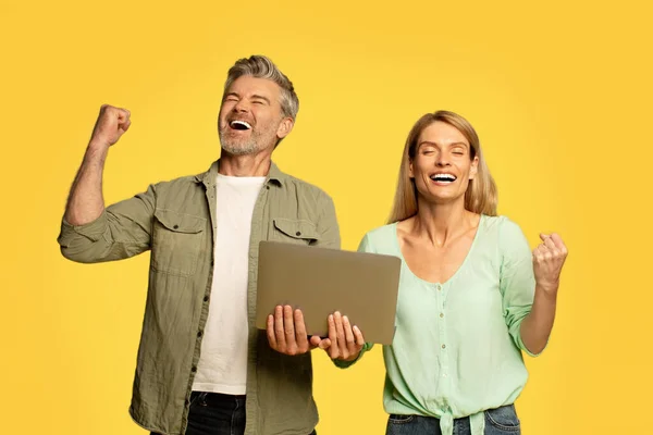 喜びに満ちた中年カップル揺れ拳を保持ラップトップコンピュータとジェスチャーはい 黄色の背景に素晴らしいニュースを祝う スタジオショット — ストック写真