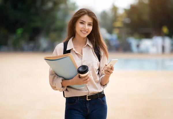 站在外面 看着相机 微笑着 拿着智能手机 咖啡和课本摆姿势的学生黑发女士 在户外向快乐的现代大学女学生开枪 — 图库照片