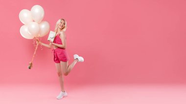 Doğum gününü kutlayan mutlu bir kadın. Elinde bir sürü balon ve hediye paketi var. Pembe arka planda duruyor.