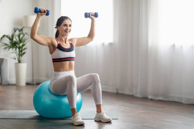 Ev antrenmanı. İyi motive olmuş, gülümseyen, beyaz spor giysili güzel esmer kadın büyük mavi bir topun üzerinde oturuyor, dambılları kaldırıyor, fotokopi alanına bakıyor, iç mekan,