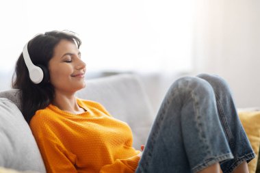 Stres atmak. Rahat, huzurlu, mutlu, günlük kıyafetler giyen, kanepeye oturmuş, kablosuz stereo kulaklık takmış, evde, yan görüşlü, kapalı gözlerle müzik dinleyen,