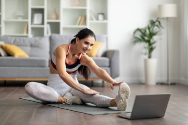 Çevrimiçi egzersiz programları. Pozitif esmer, uzun saçlı, spor kıyafetli, yoga minderinde bacaklarını esneten, evde egzersiz yapan, dizüstü bilgisayar kullanan, fitness videosu izleyen, mekanı kopyalayan bir bayan.