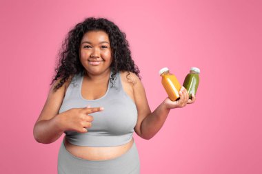 Afro-Afrikalı pozitif kadın spor kıyafetlerini tutuyor ve pembe arka planda izole edilmiş kavanozları veya meyve sularını işaret ediyor. Spor, diyet ve sağlık içecekleri için tavsiye vitaminleri