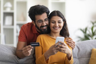 Mobil Alışveriş. Evde akıllı telefon ve kredi kartı kullanan mutlu Hintli çift oturma odasında otururken internetten ödeme yapan ve bankacılık başvurularına göz gezdiren Doğu Yakalı eşler.