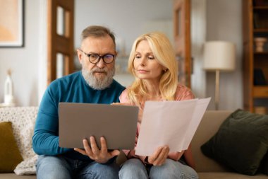 Ev Kağıtları Sayısı. Ciddi, olgun eşler dizüstü bilgisayarla birlikte evrak işleriyle uğraşıyorlar, faturaları ve yazışmaları okuyorlar Modern Ev İçişleri 'nde koltukta oturuyorlar. Gözlüklü koca.