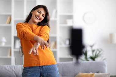 Blog konsepti. Evden yayılan rahat kıyafetler giyen güzel, uzun saçlı, doğulu bayan tripoda akıllı telefon seti kullanıyor. Ünlü nüfuzlu bayan blog kaydediyor