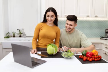 Mutlu, genç bir çift mutfak masasının başında duruyor. Evde taze organik sebzelerle dolu. Laptop kullanıyorlar.