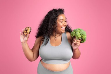 Spor kıyafetleri giymiş, brokoli yiyen, elinde donut tutan, pembe stüdyo arka planında poz veren neşeli siyah tombul kadın. Vücut bakımı, diyet ve uygun besin seçimi veya sağlıksız yiyecek, tatlı