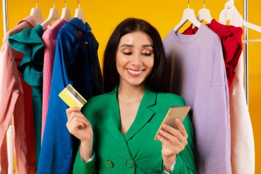 Heyecanlı genç bayan cep telefonu ve kredi kartıyla alışveriş yapıyor. Moda kıyafetlerle giyim mağazasının yanında duruyor. Sarı arka planda parlak yaz kıyafetleri sipariş ediyor.