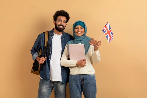 留学海外 兴高采烈的年轻阿拉伯学生 背着英国国旗的背包 快乐地笑着 一边在希吉布展示联盟杰克 一边翻阅米色工作室的背景 — 图库照片