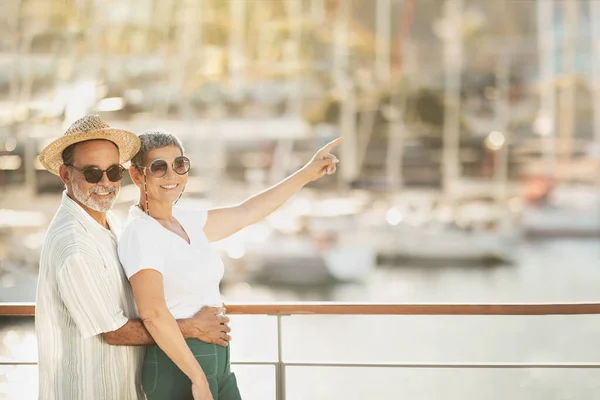 喜びに満ちた熟女観光客海のマリーナで豪華帆船を背景に指を抱きかかえて指差し 屋外に立ってカメラで微笑むカップル テキストのための空スペース — ストック写真