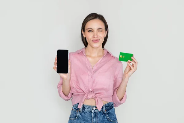 黒の空白の画面と銀行カード 素敵なモバイルアプリを表示したり 素晴らしいオファー グレーの背景 モックアップをお勧めしますと現代の携帯電話を示すカジュアルで陽気な美しい若い女性 仮想銀行 — ストック写真