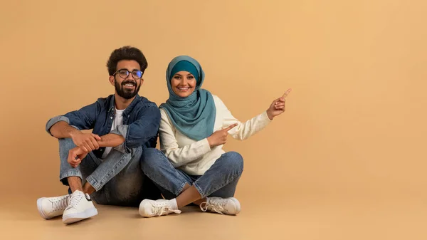 これを確認しろ コピースペースで脇を指す若いアラビア語の配偶者 あなたの広告やオファーのための無料の場所を示すヒジャブでイスラム教徒の男と女 ベージュの背景に一緒に座って パノラマ — ストック写真
