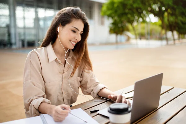 Eラーニング ノートパソコンを介して幸せなヒスパニック系の学生の女性のビデオ通話 テーブルの屋外でイヤホンを着てオンライン講義を見て学ぶ コンピュータで勉強する学習者の女性 サイドビュー — ストック写真