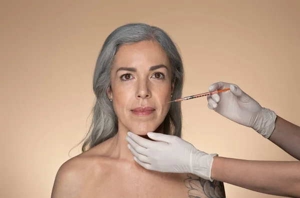 迷人的老年灰发女人在鼻唇褶皱处注射化妆品 站在米色工作室的背景上 透明质酸注射液治疗面部更新 — 图库照片