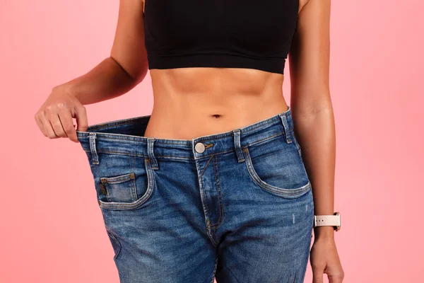 成功的饮食观念 穿着超大号牛仔裤的年轻女性展示了瘦小 无法辨认的大块头女性与体重减轻后相比的效果 她们站在粉色的背景上 — 图库照片