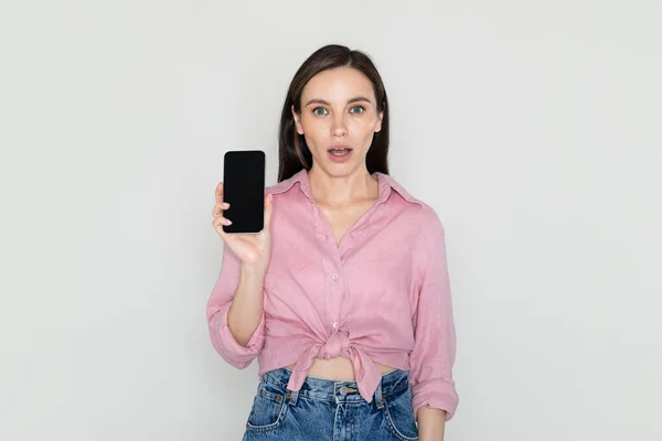 素晴らしいオンラインオファー ショックを受けたかなり若い女性を着てカジュアル服を示す携帯電話ブラックブランクスクリーン グレーの背景に隔離された モックアップ コピースペース — ストック写真