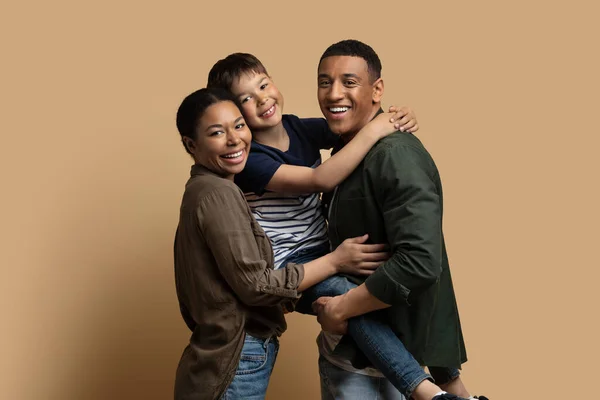 幸せな若いです両親を保持彼らのかわいい息子と笑顔でカメラ の肖像画の美しい陽気な多人種家族の3とともに子供はベージュの背景の上に一緒にポーズ コピースペース — ストック写真