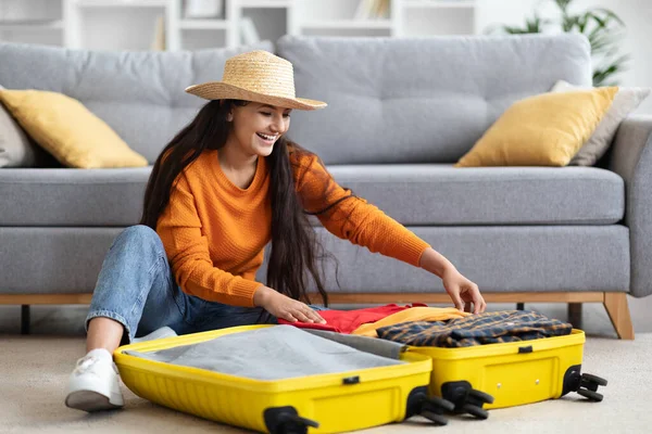 stock image Excited cheerful pretty millennial hindu woman in casual outfit and wicker hat sitting on floor in living-room, packing suitcase luggage at home, going vacation, summer trip, copy space