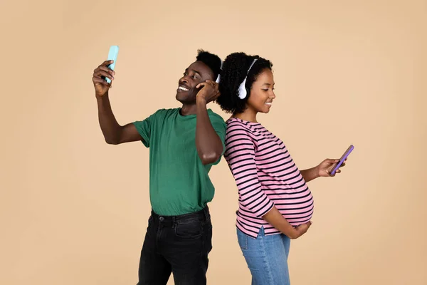 ヘッドフォンで大きな腹を持つスマートフォンや妊婦と幸せな若いアフリカ系アメリカ人男性は ベージュのスタジオの背景に隔離された自撮りを取ります 赤ちゃん 写真や楽しみ 家族を期待するためのアプリ — ストック写真