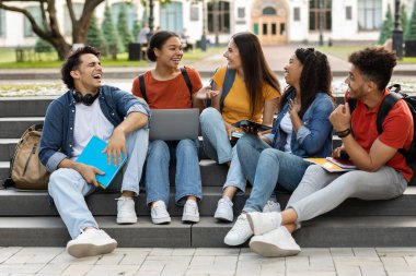 Kampüs hayatı. Neşeli çoklu etnik üniversite öğrencileri dışarıda dersler arasında dinleniyor. Laptop kullanan, sohbet eden, gülen, merdivenlerde oturan, boş alan olan mutlu erkek ve kadınlar.