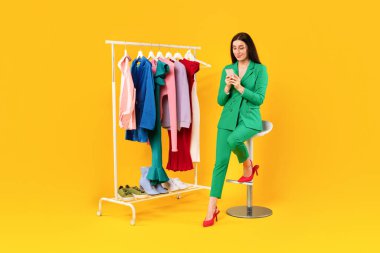 E-ticaret ve perakende. Giysileri internetten alan memnun bir bayan cep telefonuyla sarı stüdyo arka planındaki elbise rafının yanındaki sandalyede oturuyor. Tam uzunluk