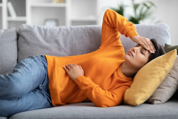 穿着休闲装的不健康的千禧年印第安妇女躺在家里的沙发上 摸着头和肚子 在经期感觉不舒服 头痛和经期抽筋 — 图库照片