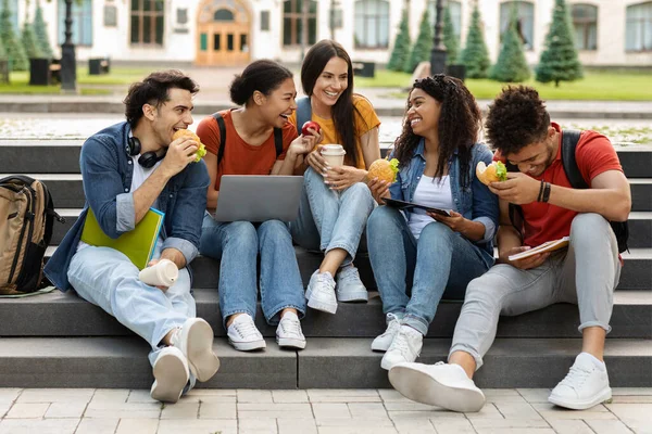 幸せな多民族学生のグループキャンパスで屋外でリラックス 陽気な正の若い友人はお菓子を食べると一緒に笑って 笑顔の男性と女性はクラスで休憩を持っています — ストック写真
