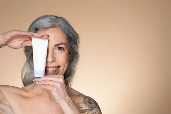 高齢女性のための美容製品 ハッピーシニア女性保持チューブともに新しい化粧品クリーム近く顔 カバー目と笑顔 ポーズ上のベージュスタジオ背景 コピースペース — ストック写真