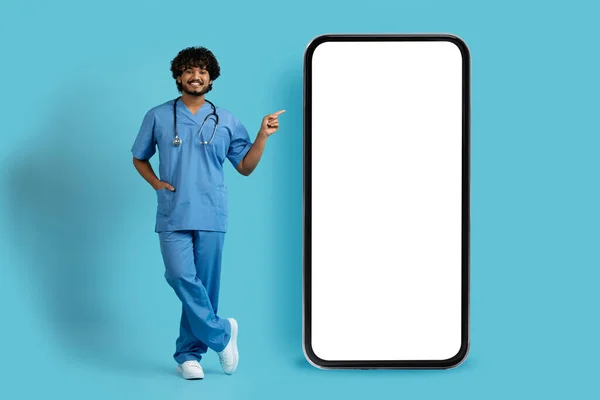 网上预约 快乐英俊 千禧年的东方人 穿着医疗工作服的医生指着装有白色空白屏幕的大手机 在蓝色的工作室背景下被隔离 模仿着 — 图库照片