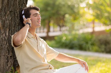 Parkta gözleri kapalı müzik dinleyen neşeli beyaz bir adam. Gardern 'de ağaçların altında oturmuş, kablosuz kulaklık takan, rahat kıyafetler giyen mutlu yakışıklı adam.