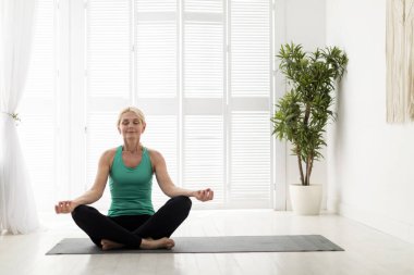 Spor Giyim 'de son sınıf güzeli evde nilüfer pozisyonunda meditasyon yapıyor, sakin olgun bayan yoga yapıyor, gözleri açık bir şekilde oturma odasındaki spor salonuna bakıyor, boşluğu kopyalıyor.