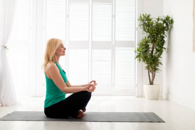 Huzurlu, olgun bir kadın evde meditasyon yapıyor, Lotus pozisyonunda gözleri kapalı oturuyor, oturma odasında meditasyon yapıyor iç mekanda iç mekanda, spor kıyafetleri içinde spor kıyafetlerini giyiyor, spor paspası üzerinde egzersiz yapıyor, fotokopi çekiyor.