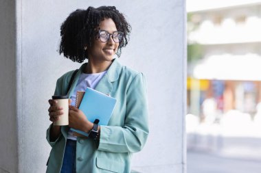 Güzel üniformalı ve gözlüklü neşeli, çekici bir genç bayan ofis binasının yanında poz veriyor, elinde not defterleri ve kağıt bardaklar, kahve içiyor, fotokopi çekiyor.