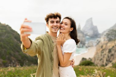 Nişanlandıktan sonra cep telefonuyla selfie çeken okyanus kıyısındaki kayalıklarda duran mutlu Avrupalı çift. Evlenme teklif ettikten sonra yüzüğünü gösteren kadın.