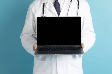 Modern bilgisayar gösteren, mavi stüdyo arka planında siyah boş ekranı olan doktor kıyafetleri, teletıp reklamı için fotokopi alanı. TeleHealth konsepti
