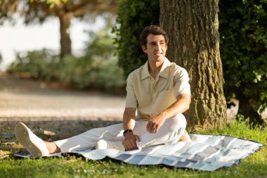 Halka açık parktaki ağaçların altında günlük kıyafetler giyen mutlu, beyaz bir adam fotokopi alanına bakıyor. Rahat adam güneşli yaz gününün tadını çıkarıyor.