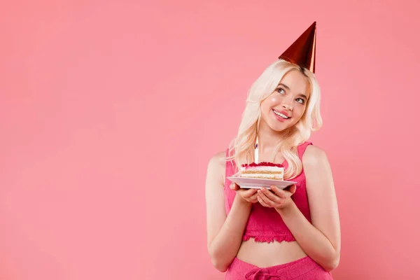 パーティーの帽子の幸せな夢の女性キャンドルとケーキのピースを保持し ピンクの背景に隔離されたコピースペースを脇に見て 誕生日と夢のための願いを作り 広告を提供し — ストック写真