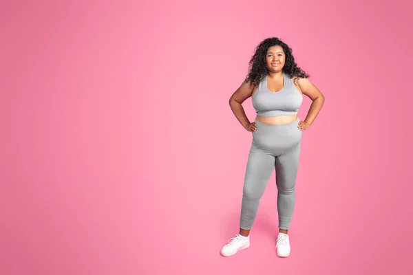 Cheerful young black oversize woman in sportswear enjoying result of workout, posing and smiling at camera on pink studio background, full length, copy space. Body care training and weight loss