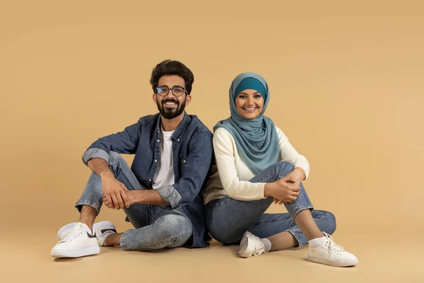 Beigeスタジオの背景に床に座っているヒジャブの幸せな若いアラビア人男性と女性 カジュアルな服で千年のイスラム教徒のカップルの肖像笑顔とカメラを見て スペースをコピー — ストック写真