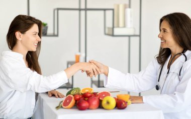 Beyaz önlüklü mutlu Avrupalı kadın beslenme uzmanı ofis içi kliniğinde genç bir bayanla el sıkışıyor. Diyet planı, tıbbi muayene, sağlık hizmetleri, kilo kaybı ve tedavi