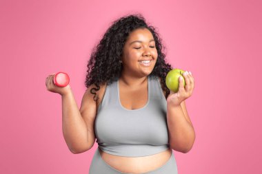 Sağlıklı beslenme konsepti. Mutlu siyah vücut pozitifliği. Elinde dambıl ve elma tutan, taze meyve ve egzersizin tadını çıkaran pembe stüdyo arka planında duran kadın.