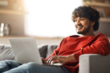 Uzaktan kumanda. Evde dizüstü bilgisayarda çalışan gülümseyen genç Hintli adam koltukta oturan ve bilgisayarını online iş için kullanan mutlu milenyum doğulu adam, uzaktaki iş fırsatlarının tadını çıkarıyor.