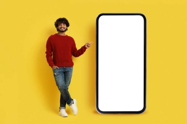 Güzel online teklif. Havalı, neşeli, yakışıklı Hintli genç adam beyaz ekranlı akıllı telefonu işaret ediyor, sarı stüdyo arka planında izole edilmiş, model, kopyalama alanı
