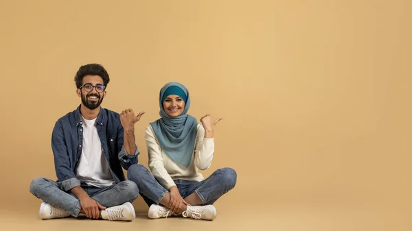 素晴らしいオファーだ ムスリムカップルの笑顔が2つの親指でコピースペースを指します 無料の場所を示すヒジャブで幸せなアラビア人男性と女性 ベージュスタジオの背景に座って パノラマ — ストック写真